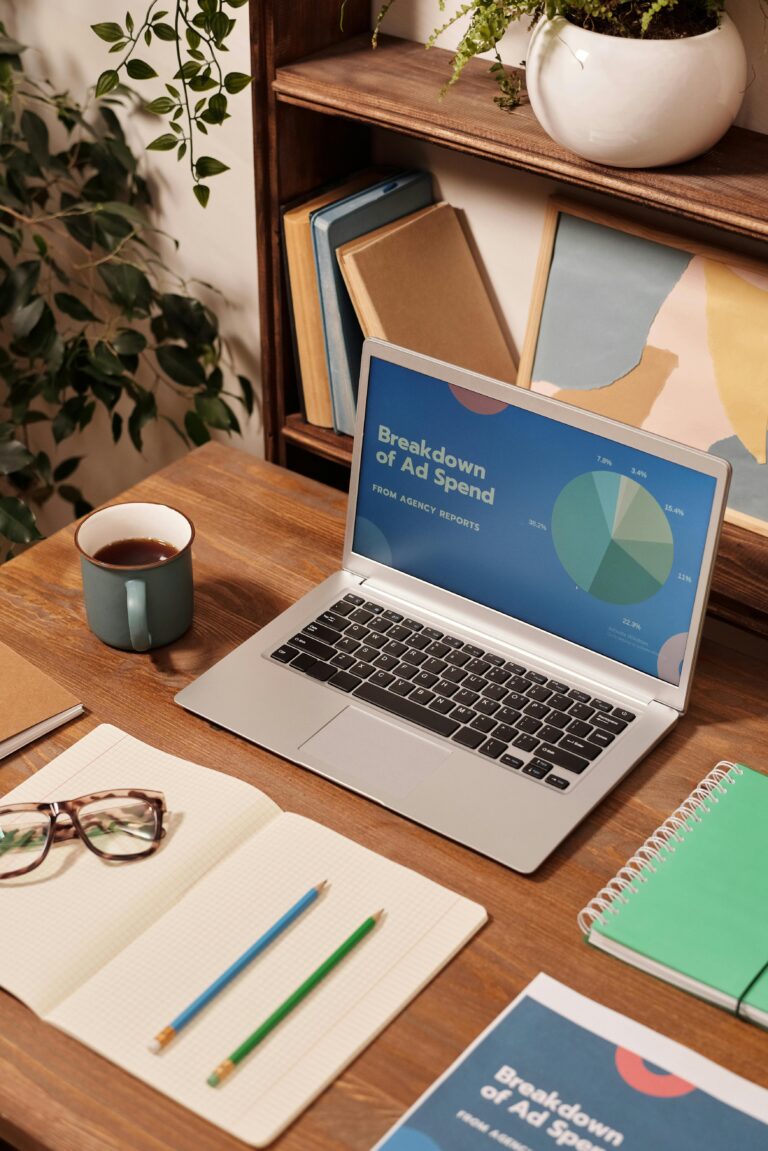 Workspace with a laptop showing an ad spend report, notebooks, and coffee cup on wooden desk.